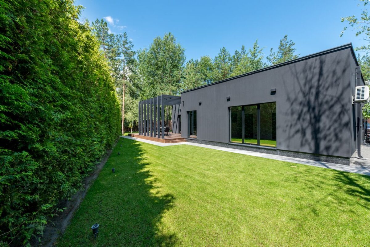 Offres Sleek black modern house with large windows and vibrant green lawn surrounded by dense trees.